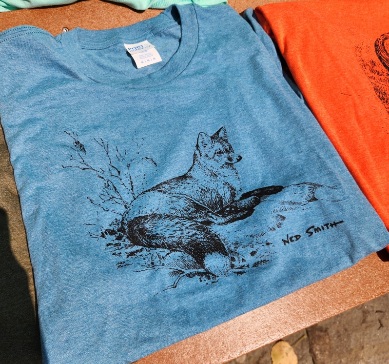 Product Description: Ned Smith Original Artwork T-Shirt each with a conservation quote on the back, and the Ned Smith Center for Nature and Art Logo on the left sleeve.</br>All proceeds go to the continuance of the legacy of Ned Smith and support The Center to provide children's camps, educational programs, owl banding, fungi programs, concerts, and art preservation.</br></br>Blue Fox ©, "All good things are wild and free" -Henry David Thoreau</br>Green Raccoon ©, "Leave the road, take the trails" -Pythagoras</br>Teal Brook Trout ©, "In nature nothing exists alone" -Rachel Carson</br>Orange Owl ©, "Conservation is a state of harmony between men and land" -Aldo Leopold</br> 923 NSC T-Shirt Assorted